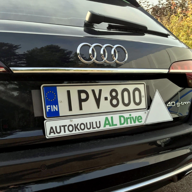 Lähikuva Autokoulu AL Driven ajo-opetusauton mustan Audin takaosasta, missä autokoulun logo, rekisterikilpi ja ajo-opetuksen ilmoittava valkoinen kolmio.