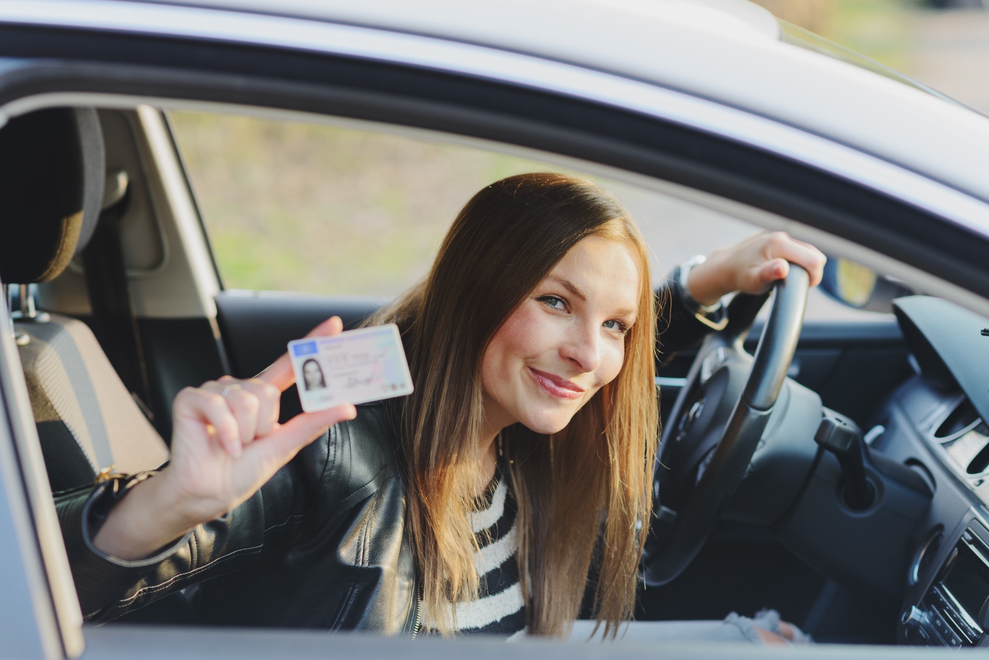 Nuori nainen istuu autossa ratin takana iloisena ja nayttaa ajokorttiiaan.