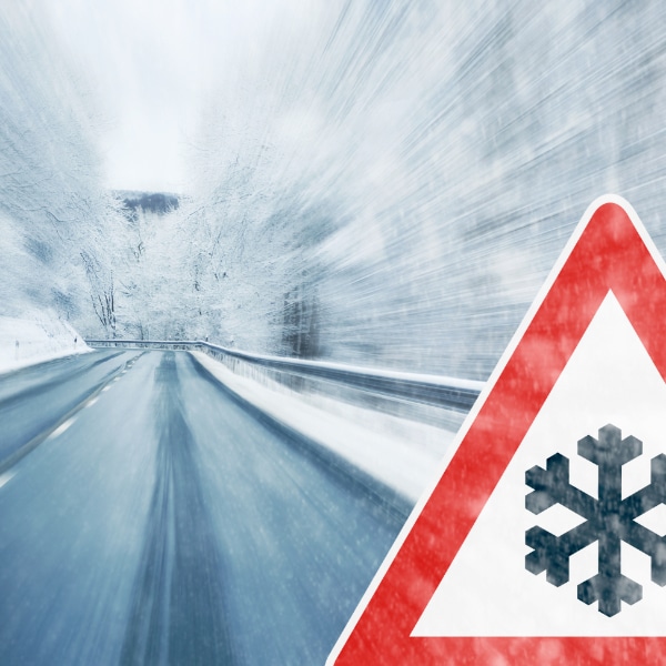 Epaterava kuva talvisesta liukkaasta tiemaisemasta, kuvan etualalla lumisateesta varoittava liikennemerkki.